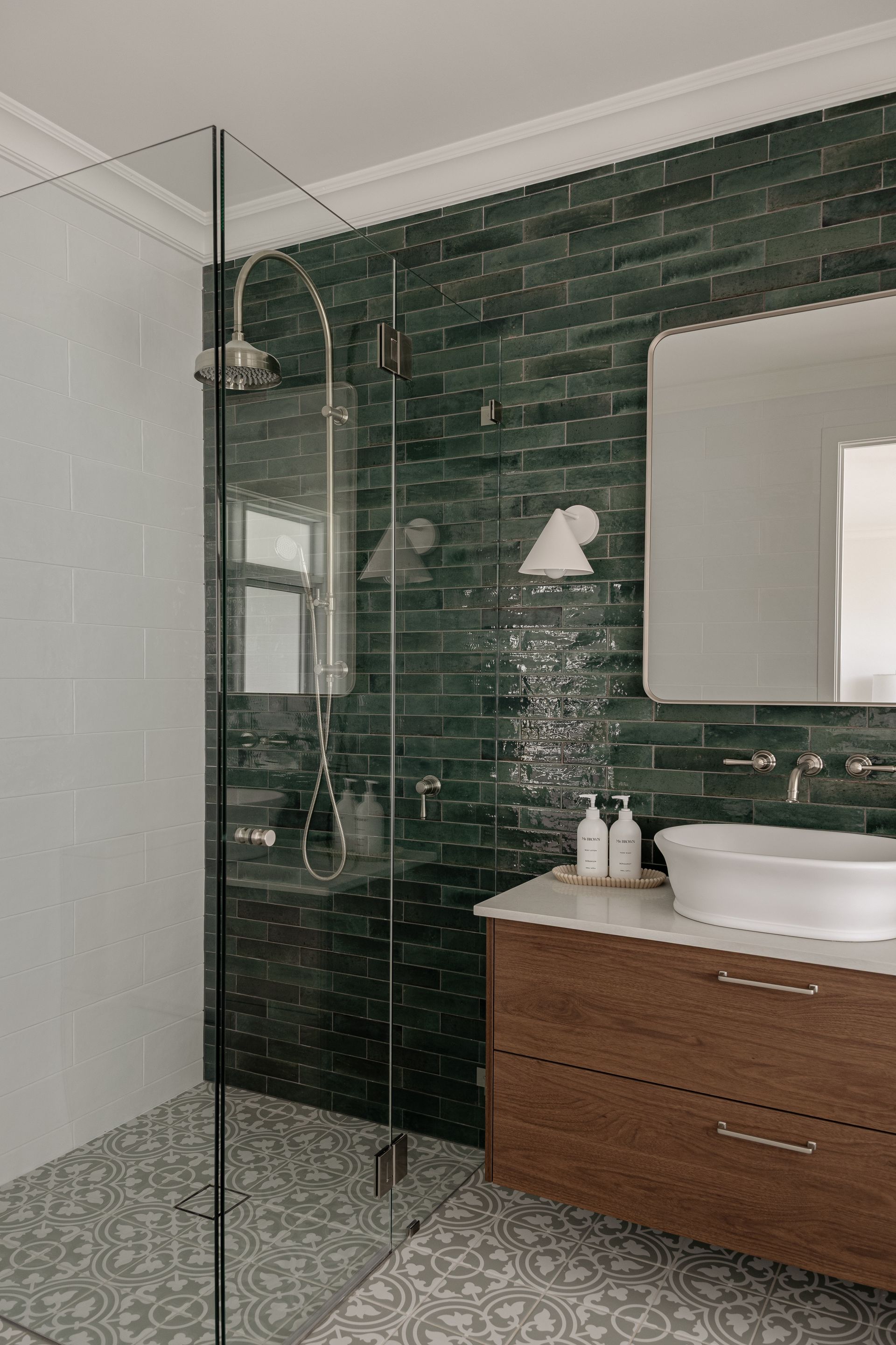 Green-tiled shower with glass enclosure. Wooden vanity with vessel sink. Patterned floor.