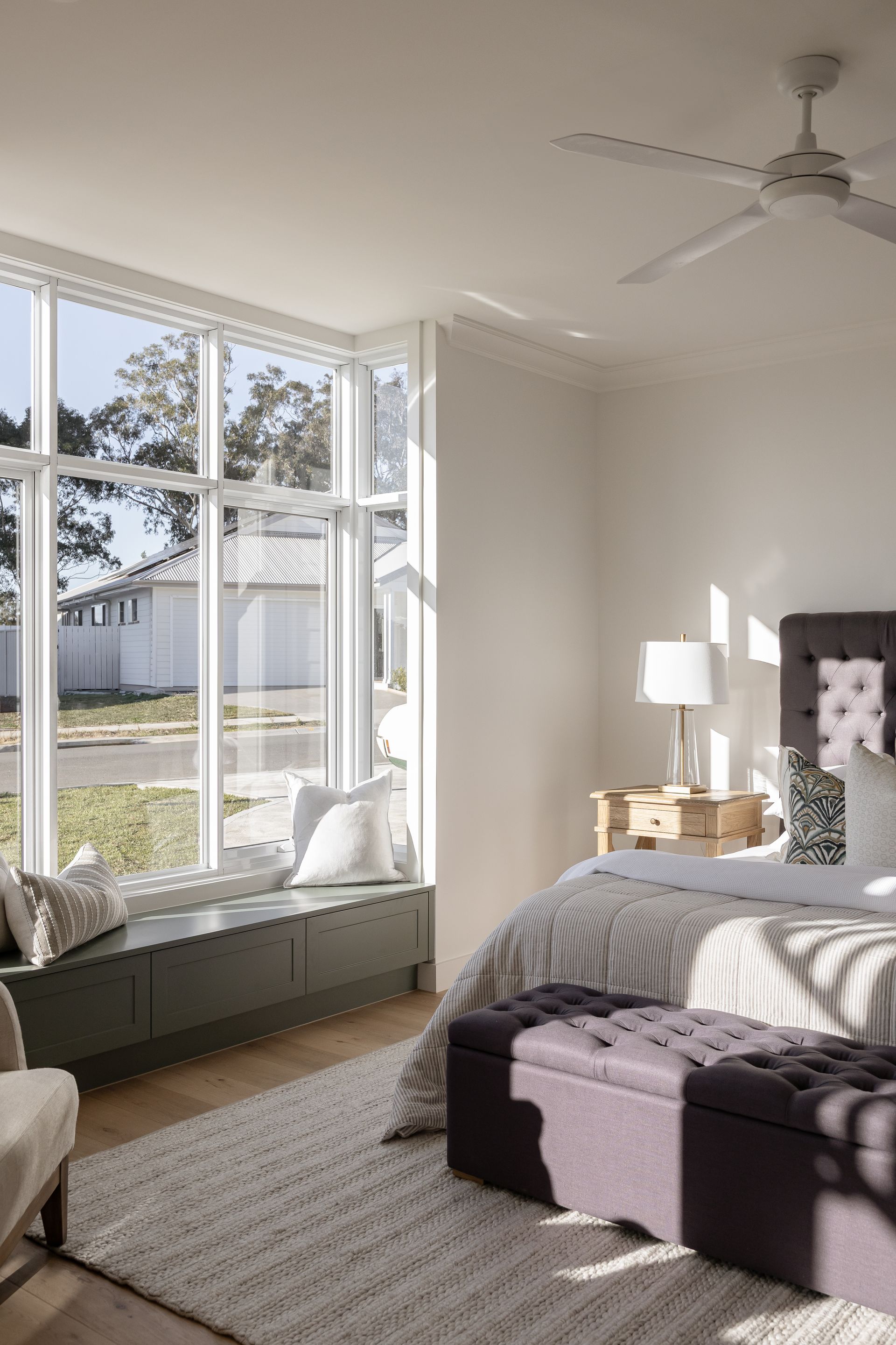 Bedroom with large windows, gray built-in bench, purple ottoman, and neutral-toned decor.
