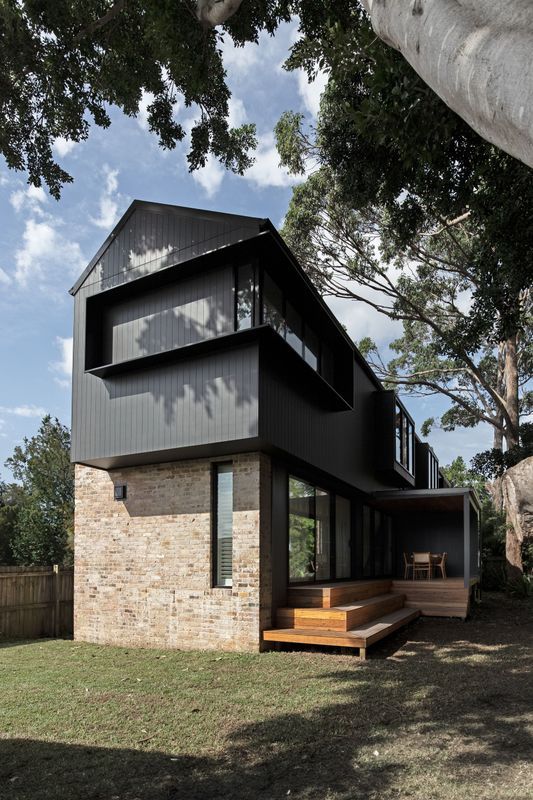 Modern house with dark wood siding on top of a brick base; wooden deck and green yard.