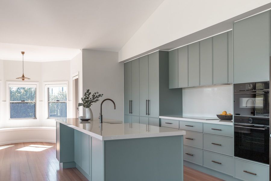 Modern kitchen with blue-green cabinets, white countertops, and a black oven. Island with a black faucet.