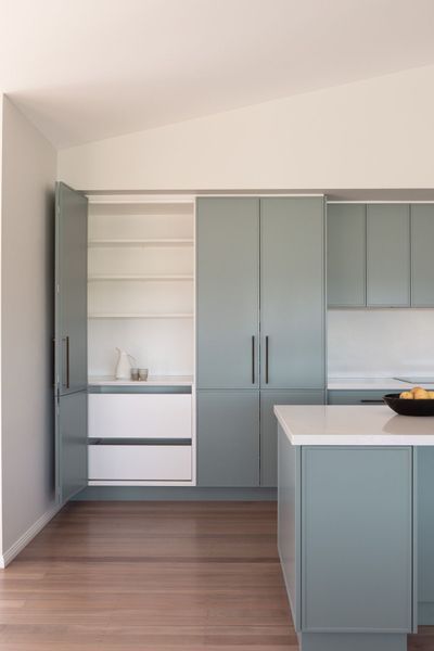 Modern kitchen with blue-green cabinets, pantry open, white countertop island, and wood floor.