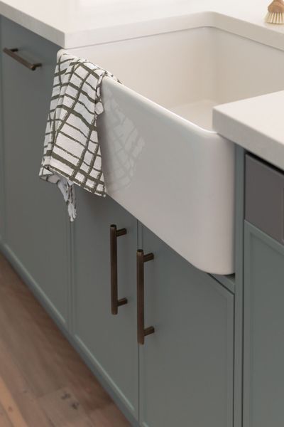 A white farmhouse sink with blue-green cabinets and dark bronze handles; a dish towel hangs on the sink.
