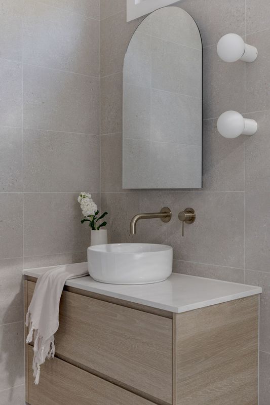 Bathroom vanity with a light wood cabinet, white countertop, and vessel sink; arched mirror, gold faucet, wall sconces.