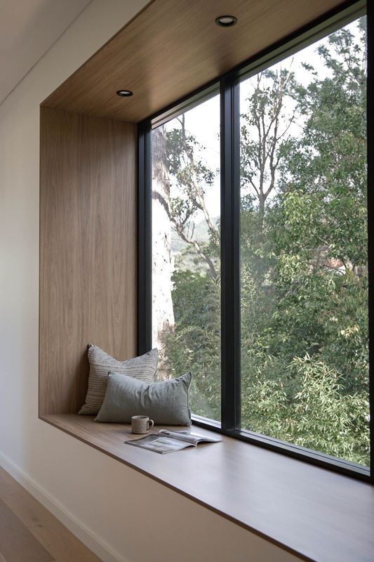 Built-in window seat with wooden frame, pillows, and a book, overlooking a green tree canopy.
