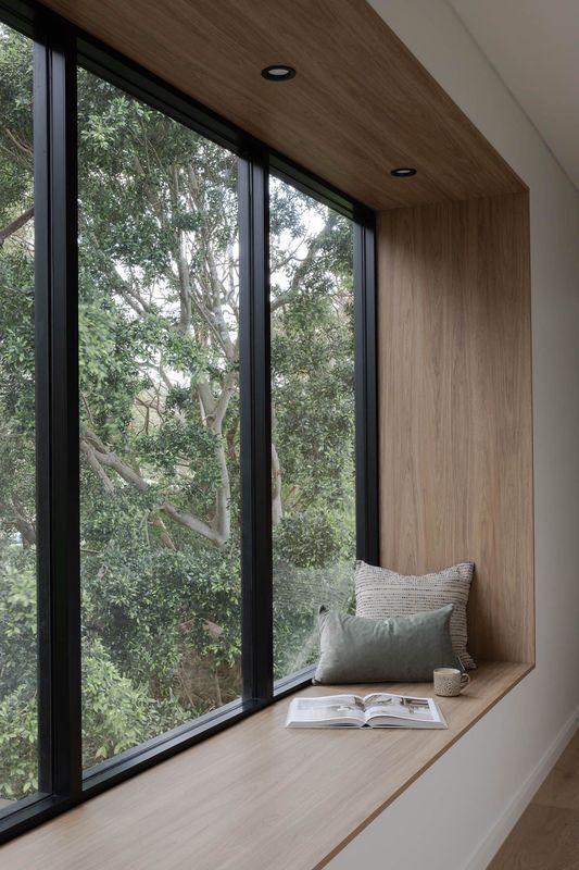 Window seat with wood paneling, black frame window overlooking lush green trees. Cushions and book rest on the seat.