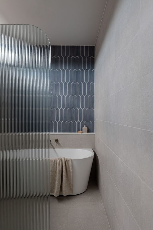 Modern bathroom with a blue tile wall, white tub, and clear, ribbed glass shower partition.