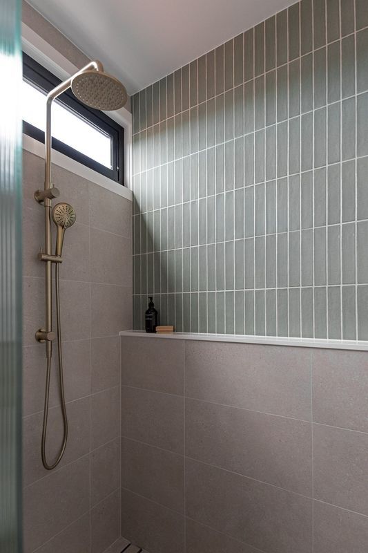 Shower with grey tiles and gold fixtures. Showerhead and handheld, shampoo bottle, and soap.