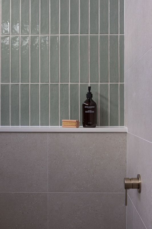Shower with light green vertical tile backsplash, gray lower tiles, shelf with soap and bottle.