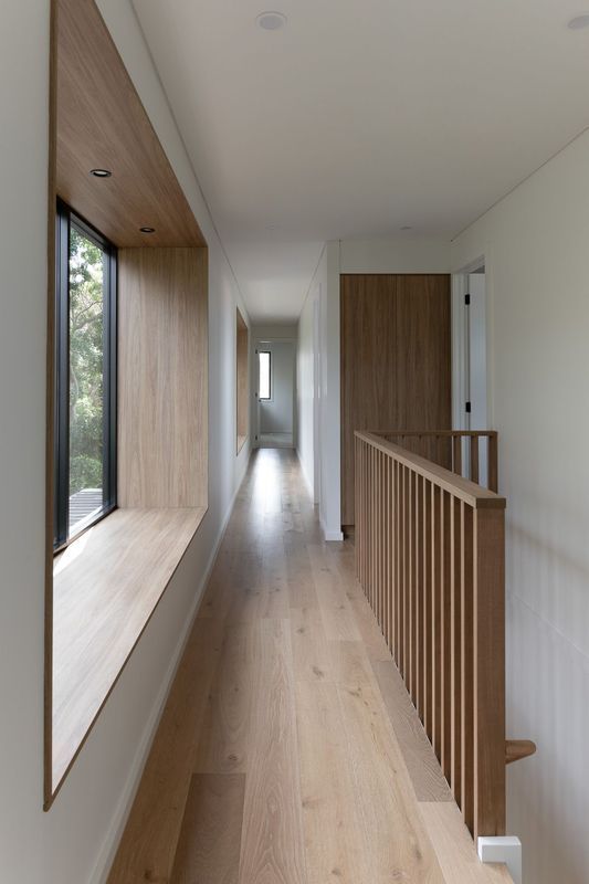 Long hallway with wooden floor, railing, and window seat, with white walls and a wooden door.