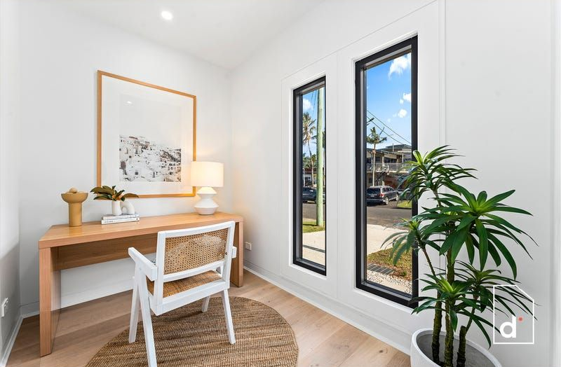 Home office with a wooden desk, woven chair, and natural light from tall windows.