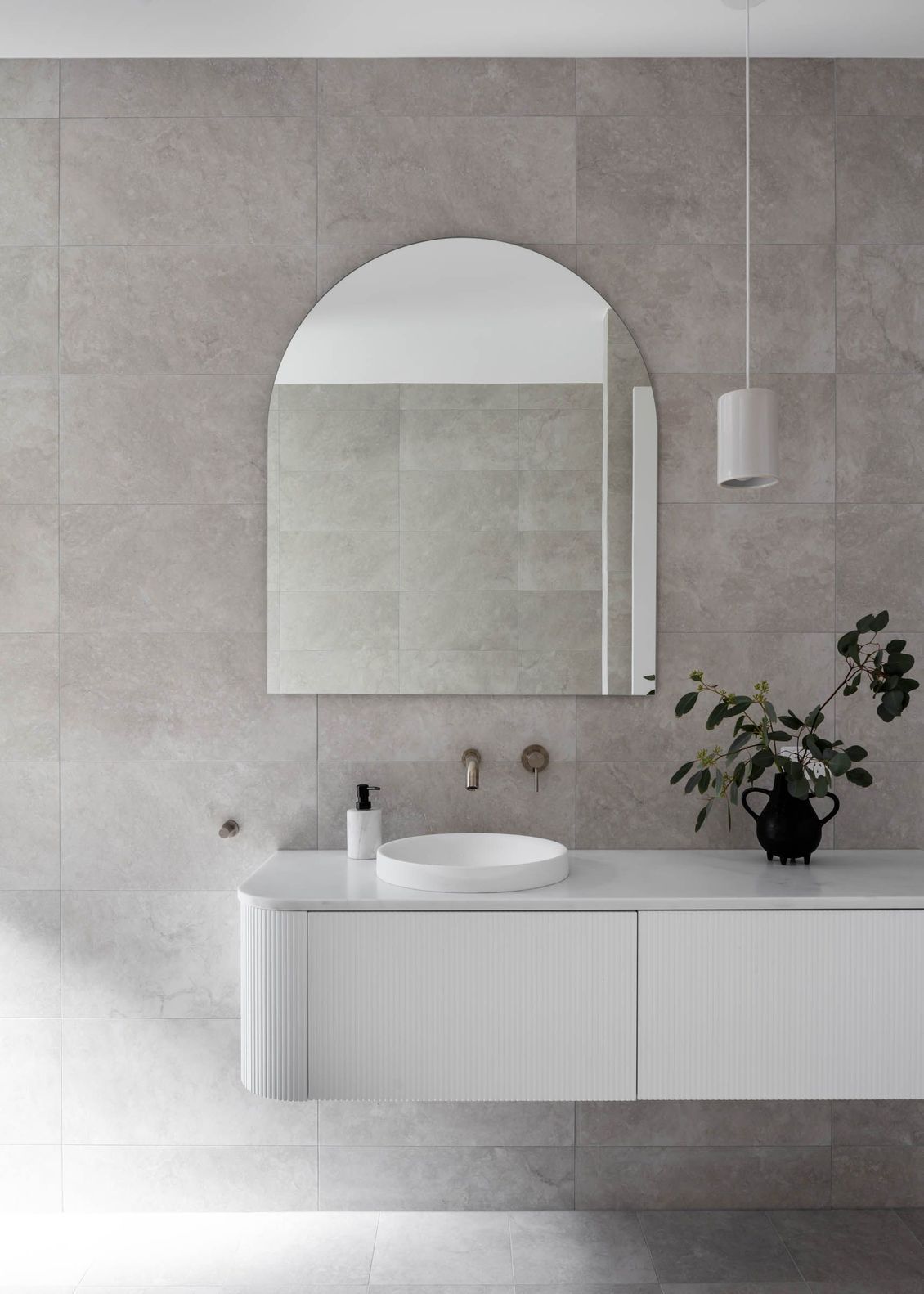 White modern bathroom with arched mirror, floating vanity, gray tiled walls, and decorative plant.