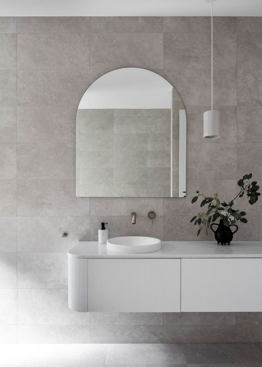 White modern bathroom with arched mirror, floating vanity, gray tiled walls, and decorative plant.
