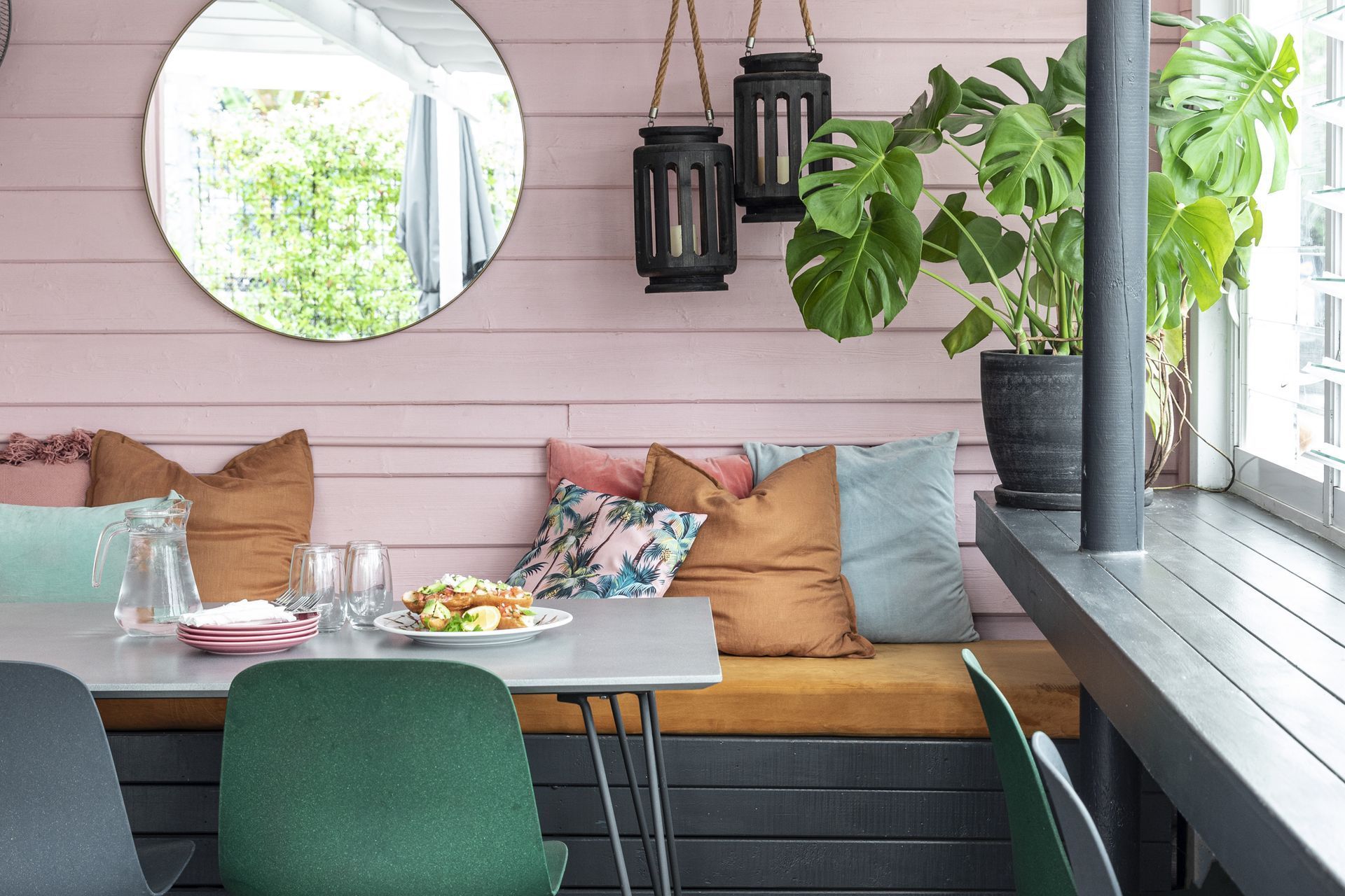 Outdoor seating area with pink wooden walls, round mirror, and Monstera plant.
