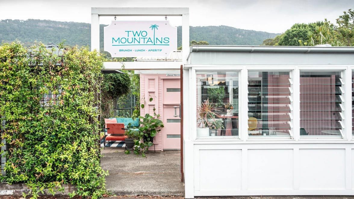 Two Mountains shop exterior; pink door, white building, sign, greenery, mountains in background.