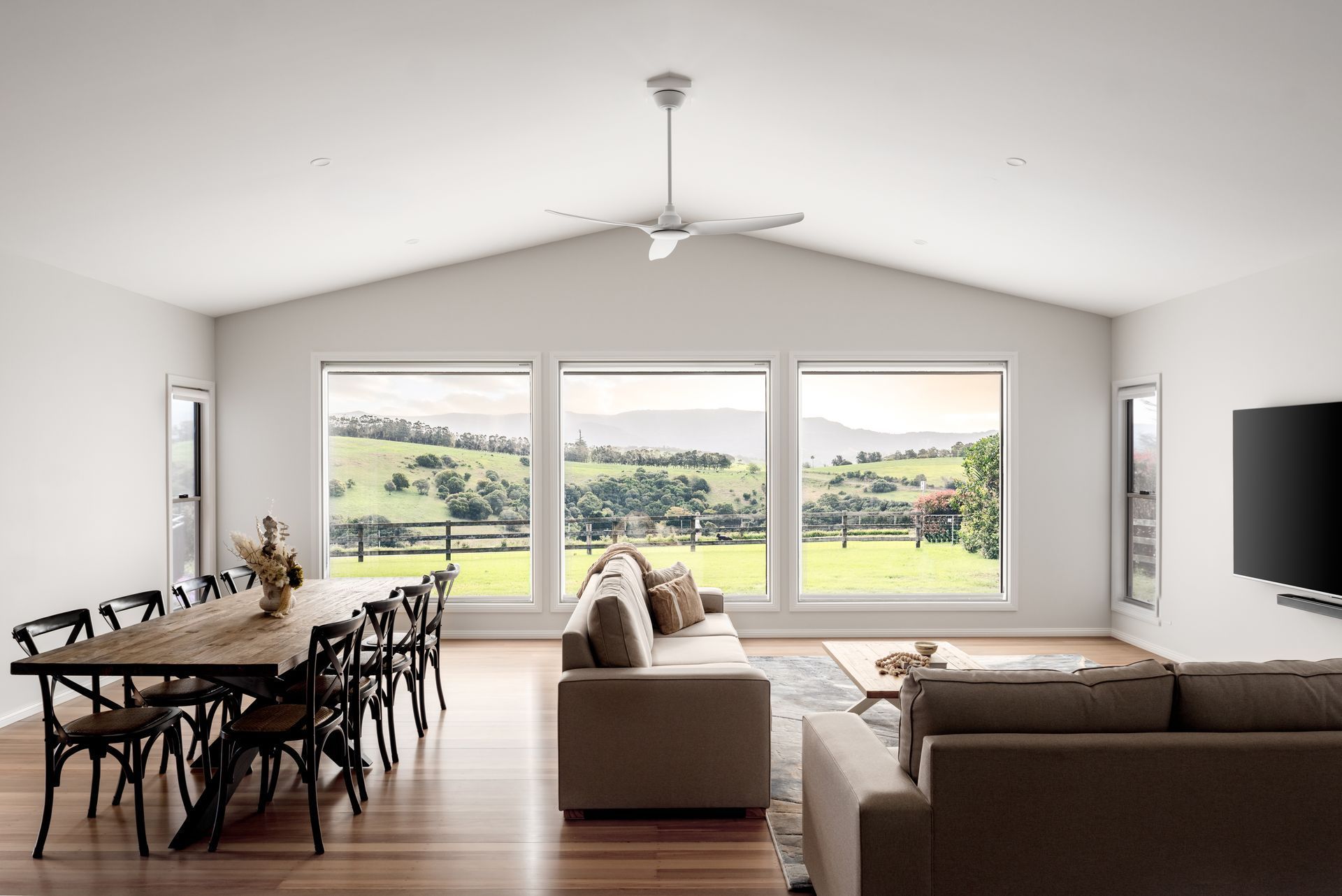 Living room with large windows offering a scenic view. Features a dining table, sofas, and a TV.