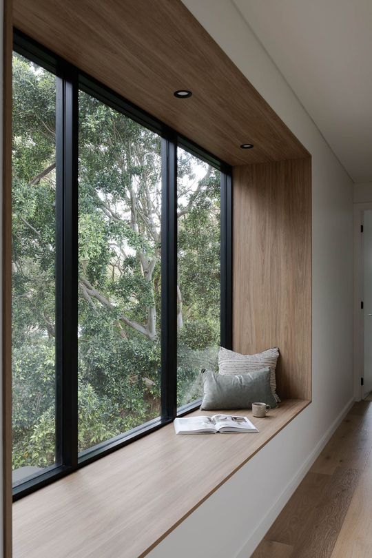 Window seat with wooden frame, overlooking green trees. Cushions, book, and cup are on the seat.