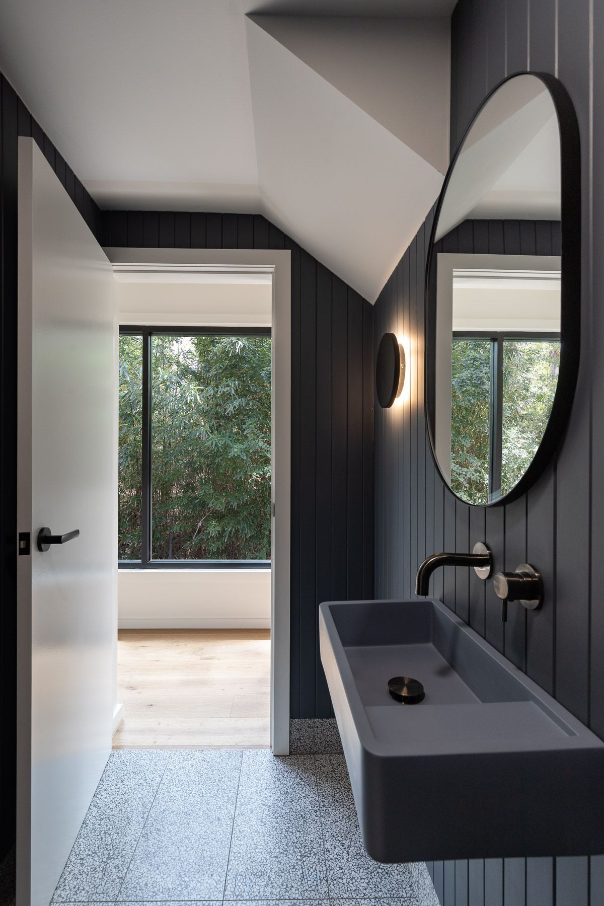 Dark gray bathroom with a floating sink, oval mirror, and vertical paneling. Doorway leads to a window with a forest view.
