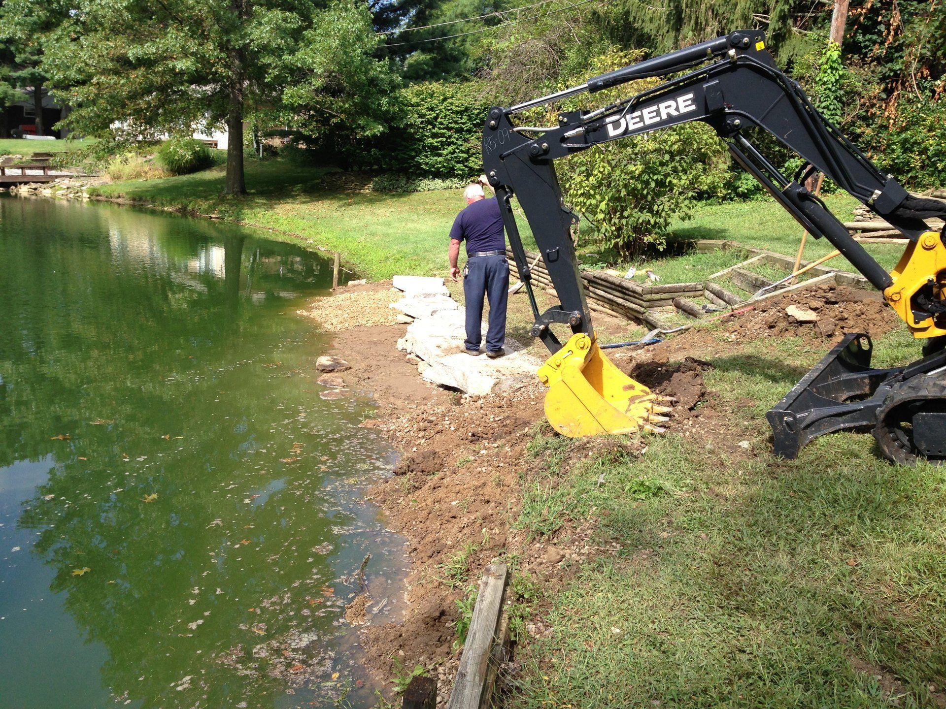 A man is standing next to a deere excavator near a lake.