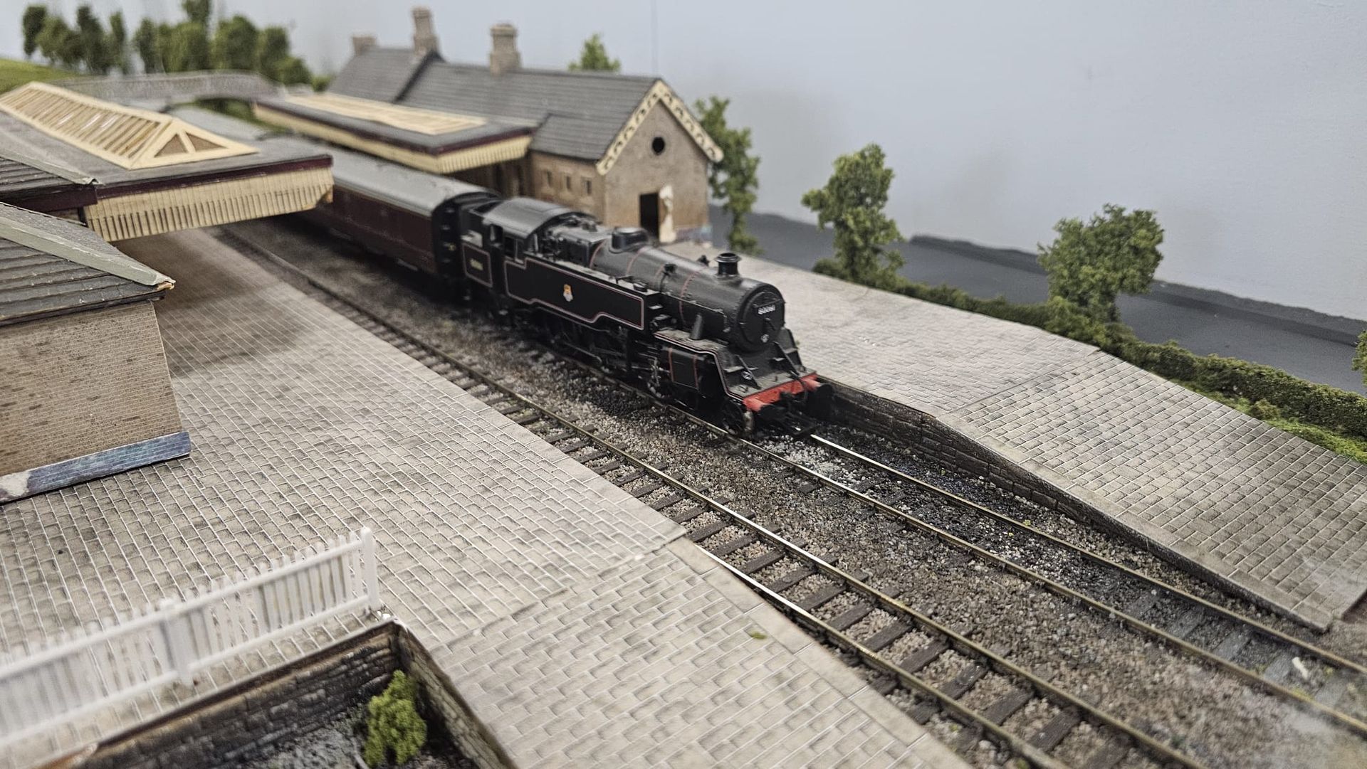 Model train entering a station with black locomotive, brown carriages, and brick buildings.