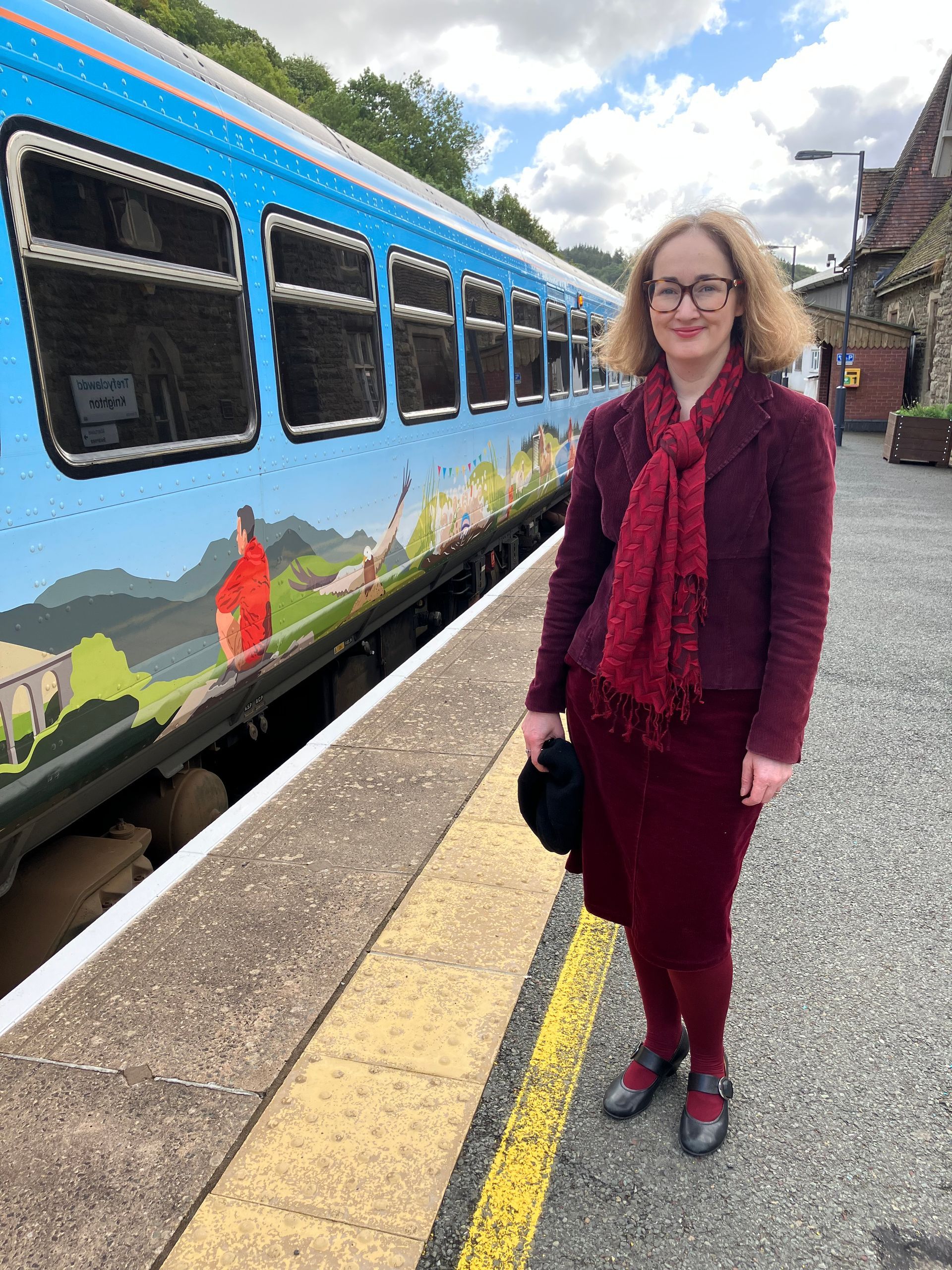 Woman in maroon attire stands on a train platform next to a blue and white train with a landscape mural.