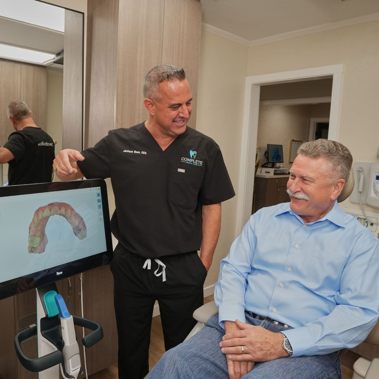 A man in a blue shirt sits in a dental chair while a man in a black shirt stands next to him