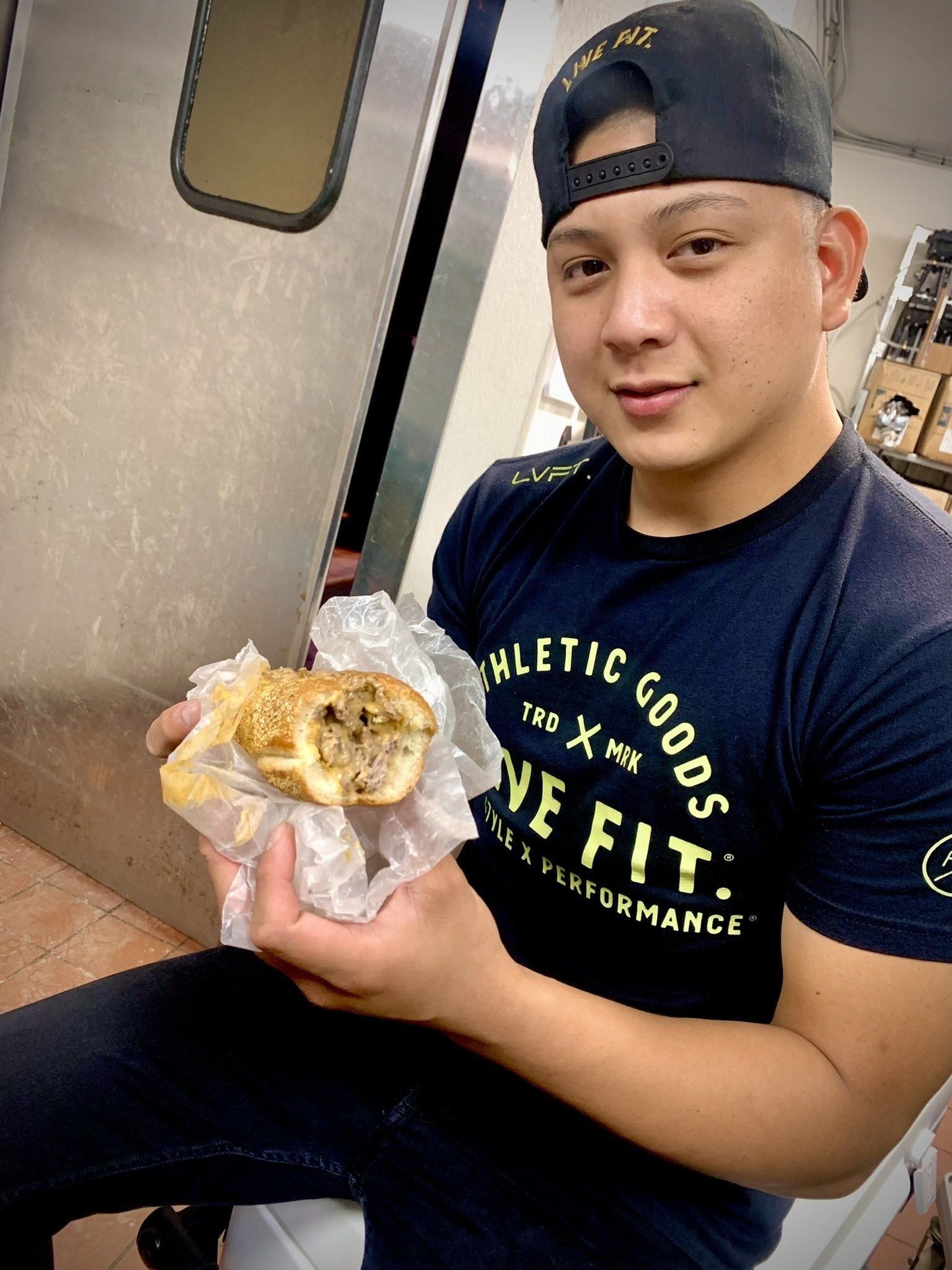 Man holding a partially eaten sandwich, smiling. Wearing a cap and a dark blue shirt in a food service area.