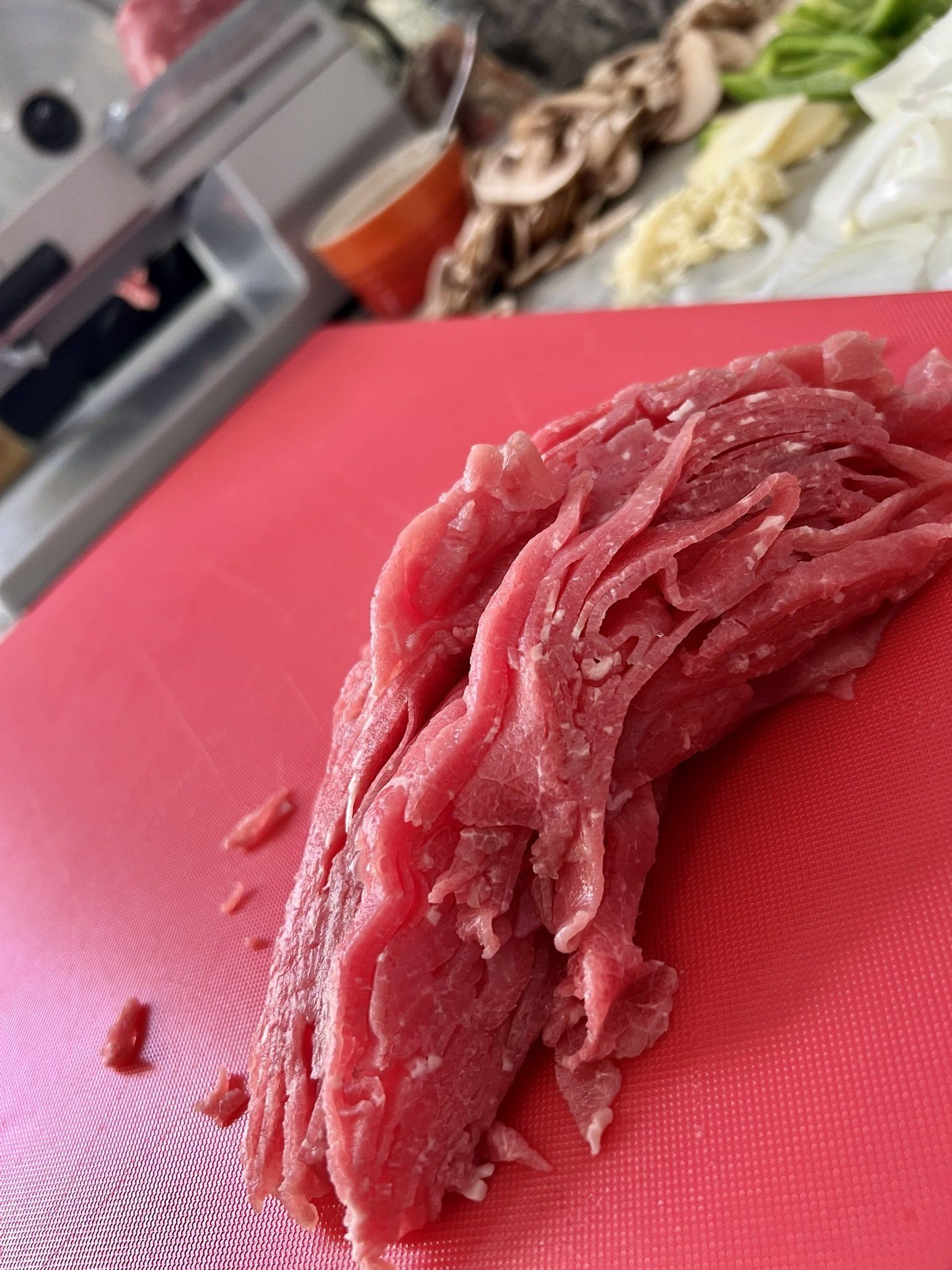 Thinly sliced raw beef on a red cutting board, with vegetables in background.
