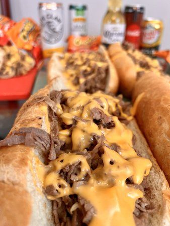 Close-up of cheesesteak sandwiches on a table, with beer, soda, and snacks in the background.