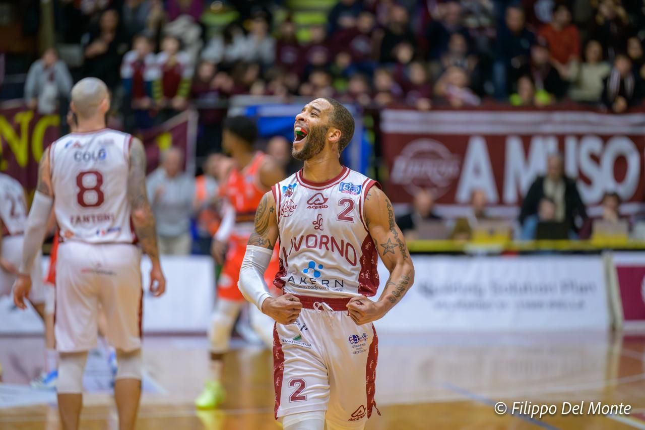 Un giocatore di basket festeggia indossando una maglia bianca con dettagli rossi e i pugni chiusi in un'arena gremita.