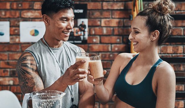 Un uomo e una donna in abiti sportivi brindano con i loro frullati, sorridendo. Ambiente da palestra.