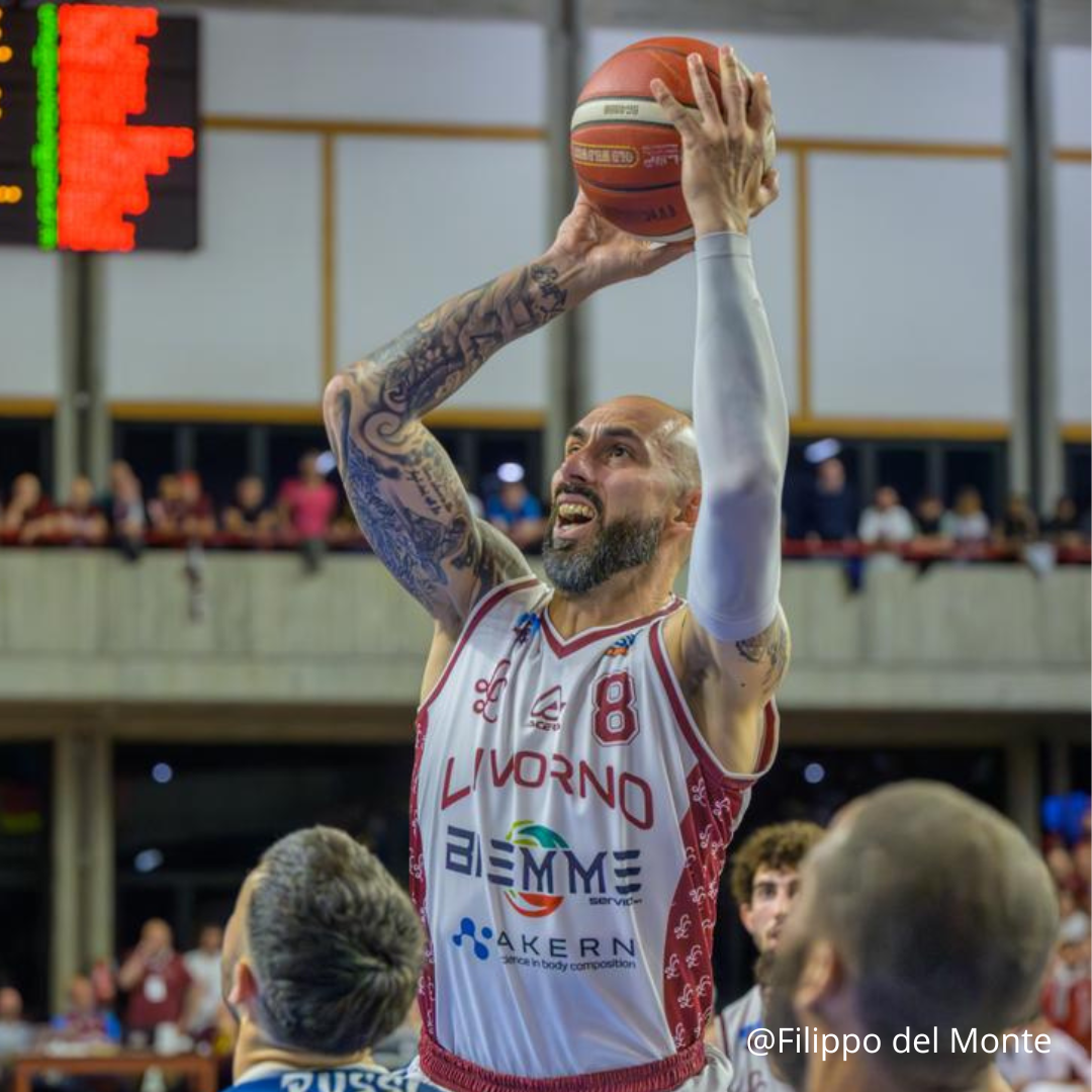Giocatore di basket che tira una palla; maglia rossa e bianca, tatuaggi, in casa, espressione concentrata.