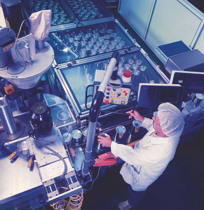 Una persona in camice da laboratorio aziona dei macchinari in una fabbrica; è visibile la linea di produzione dei contenitori.