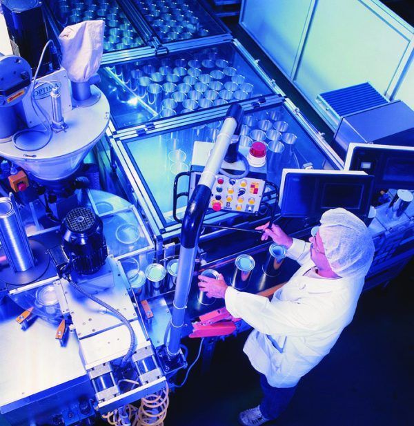 Operaio in camice da laboratorio che aziona macchinari e riempie lattine in una fabbrica illuminata da luci blu.