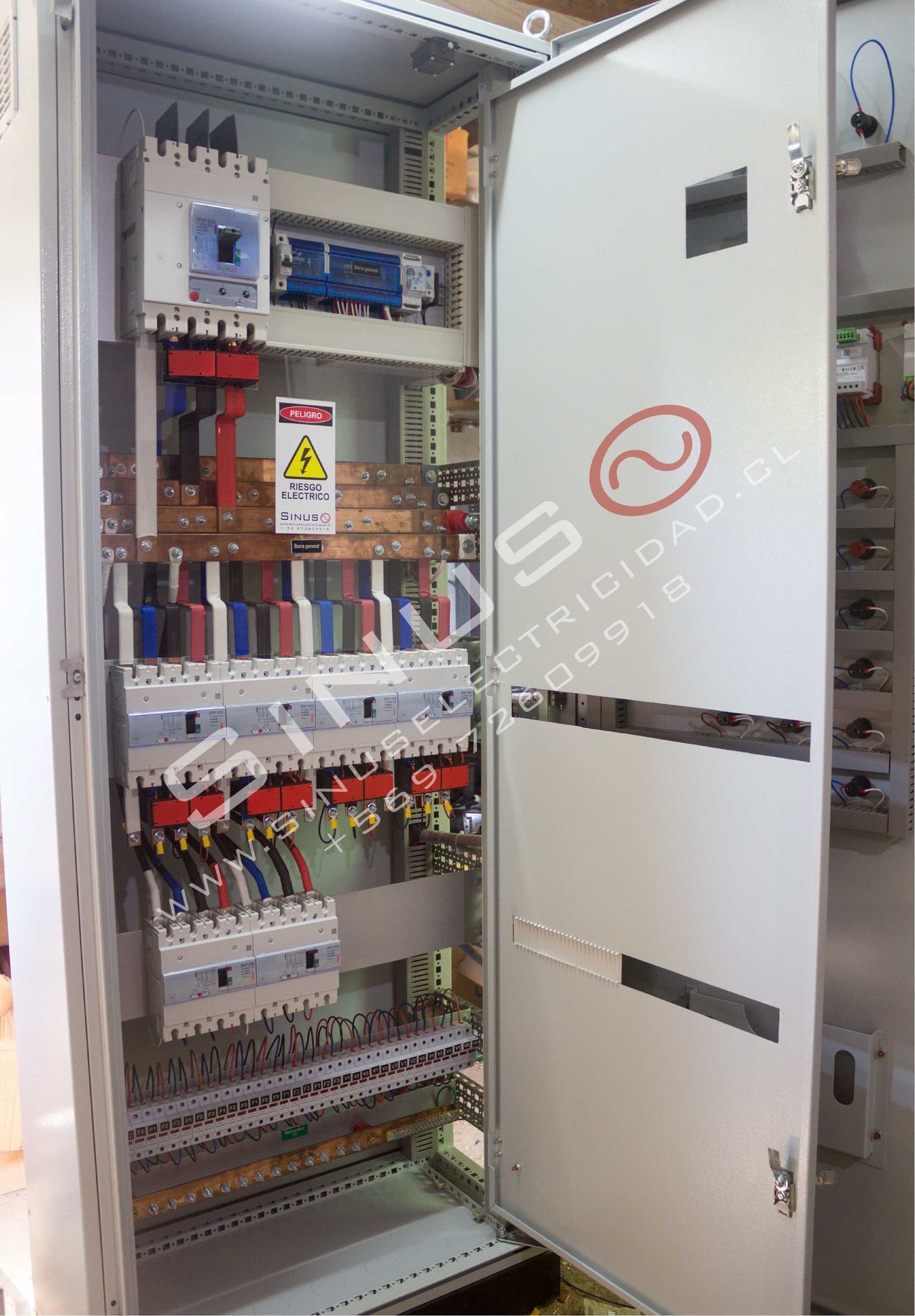 Panel de control eléctrico abierto con cableado, disyuntores y una puerta blanca en un entorno industrial.