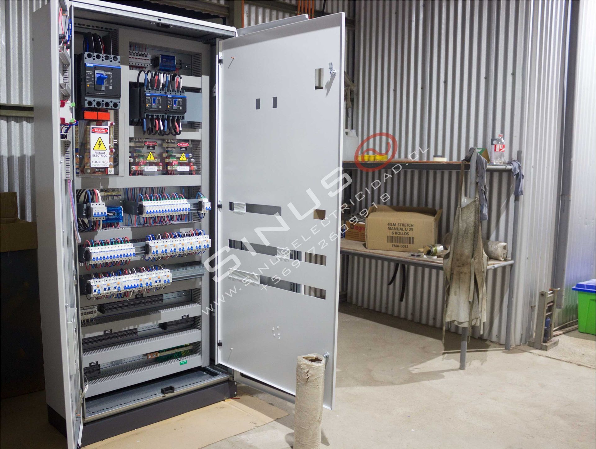 Armario de control eléctrico abierto en un taller industrial con cableado, interruptores y etiquetas a la vista.