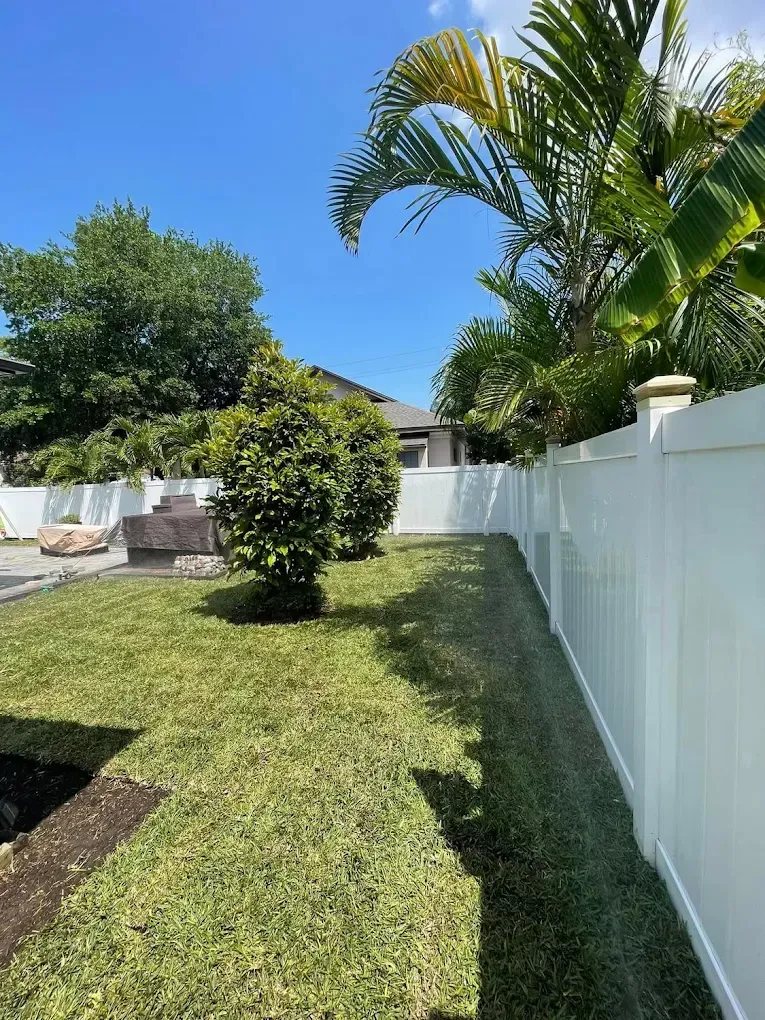 Green lawn bordered by a white fence with green trees under a blue sky.