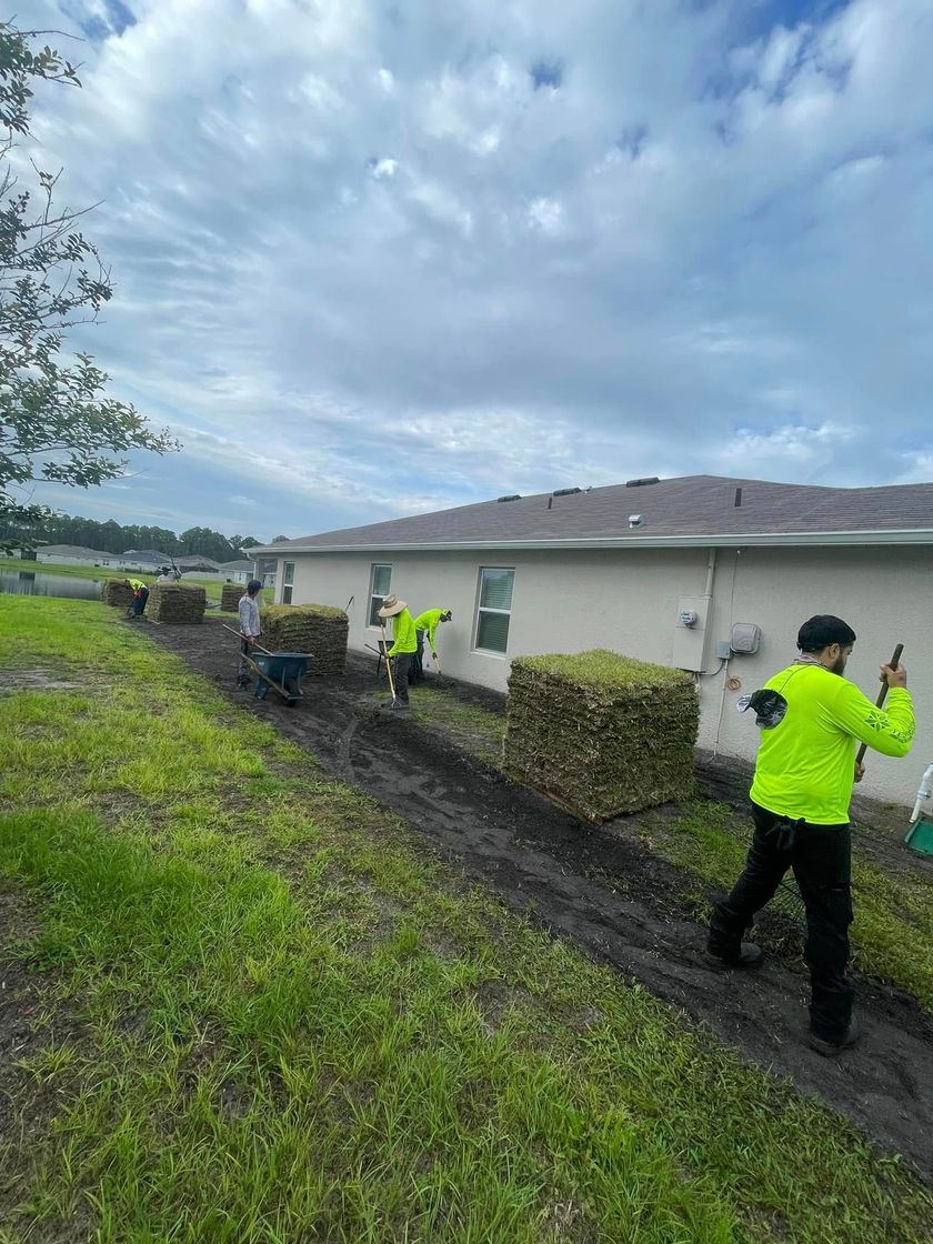 Workers laying sod along a house, wearing neon green shirts. Muddy ground, cloudy sky.
