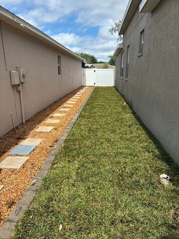 Narrow yard between two buildings; stone pathway on left, grass on right, white fence in the distance.