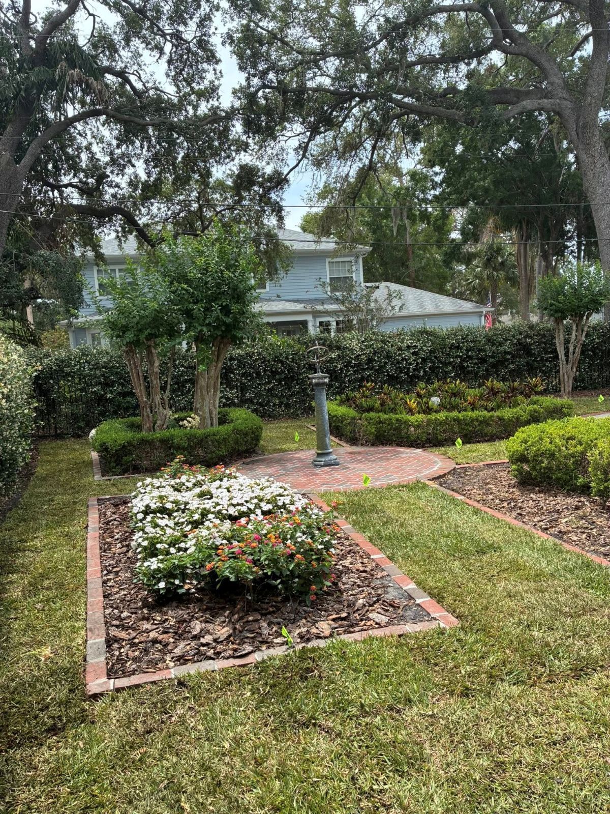 Manicured garden beds with brick borders, green bushes, and a light blue house in the background.
