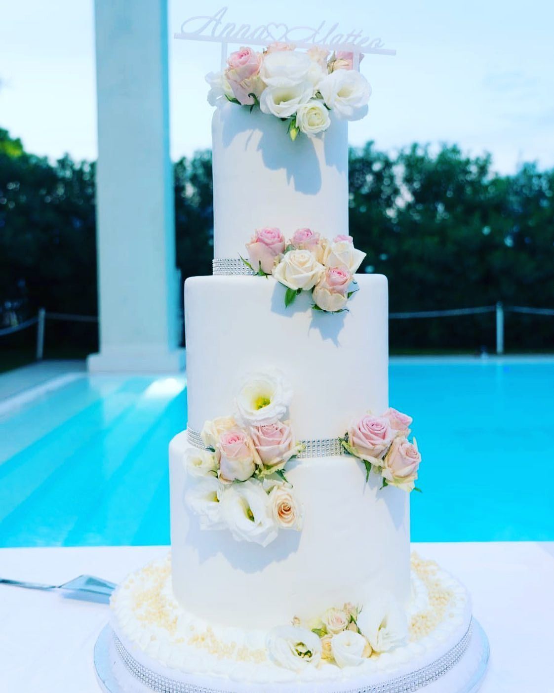 Torta nuziale bianca a tre piani decorata con fiori rosa e bianchi, a bordo piscina.
