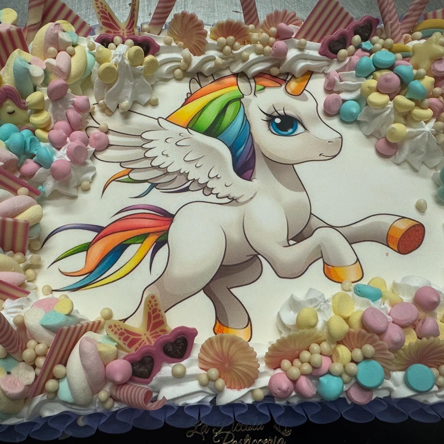 Torta unicorno con criniera e coda arcobaleno, ali, circondata da caramelle e dolciumi, glassa bianca.