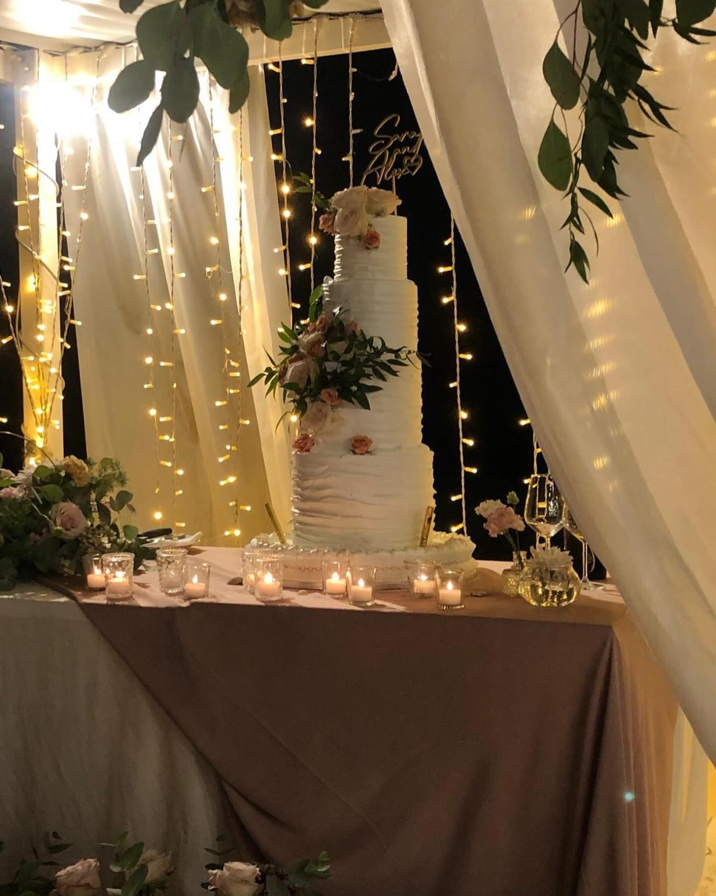 Torta nuziale su un tavolo ricoperto di stoffa, decorato con fiori e candele, sotto lucine.