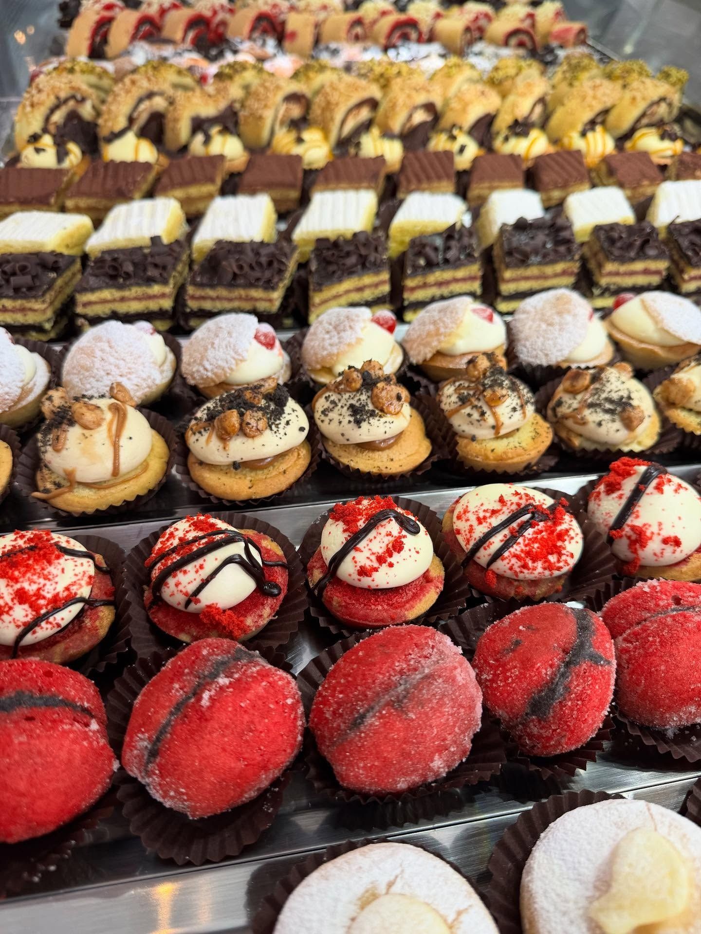 File di pasticcini assortiti, per lo più in formato boccone, con colori e guarnizioni diversi, esposti in una vetrina di vetro.