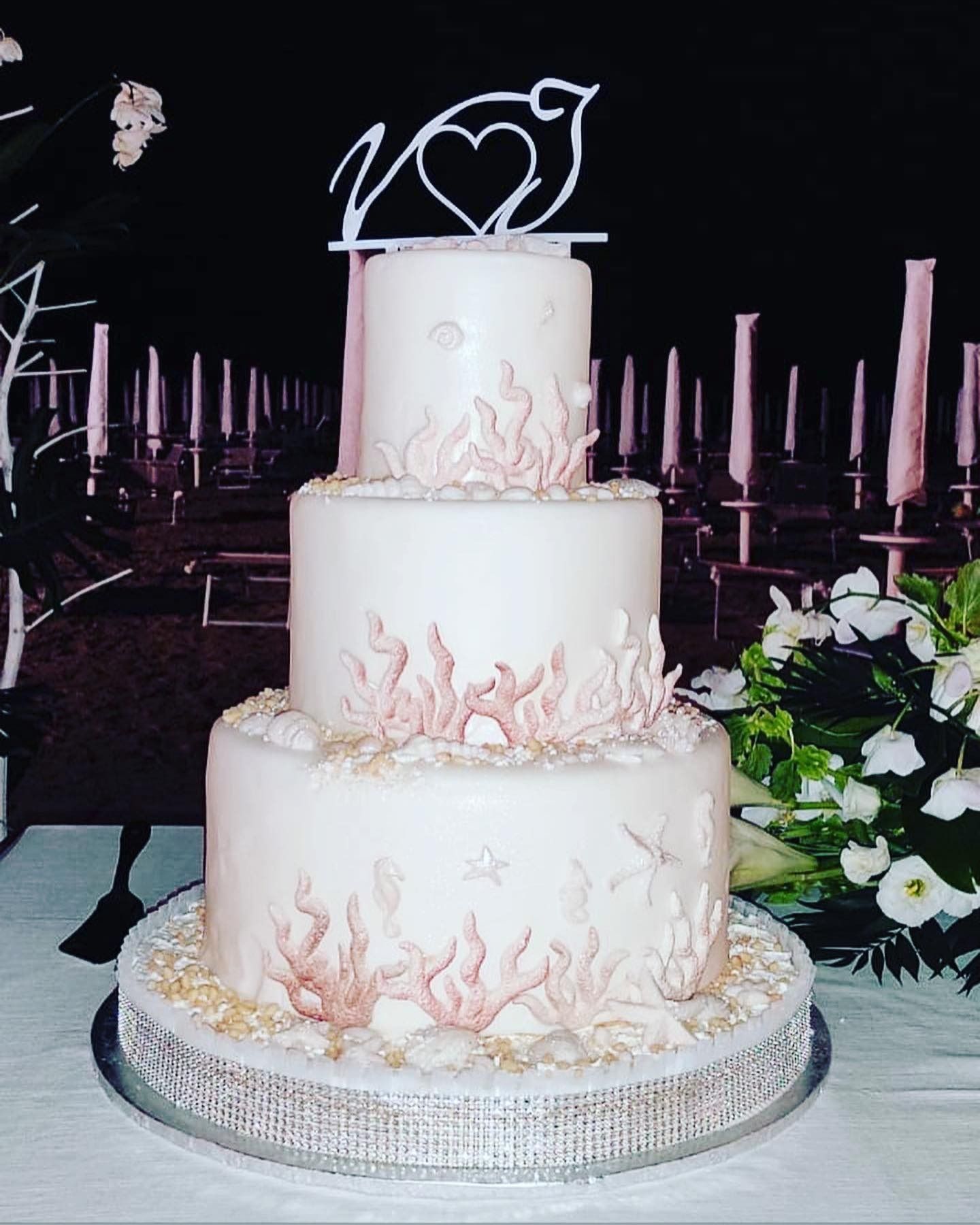 Torta nuziale bianca a tre piani con dettagli in corallo e un cuore a forma di uccello in cima, allestita su una spiaggia.