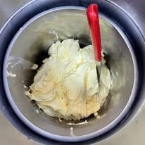 Sostanza cremosa, di colore giallo pallido, in un contenitore di acciaio inossidabile con una spatola rossa.