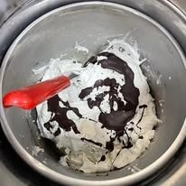 Gelato con vortici di cioccolato in una gelatiera di metallo, mescolato con una spatola rossa.