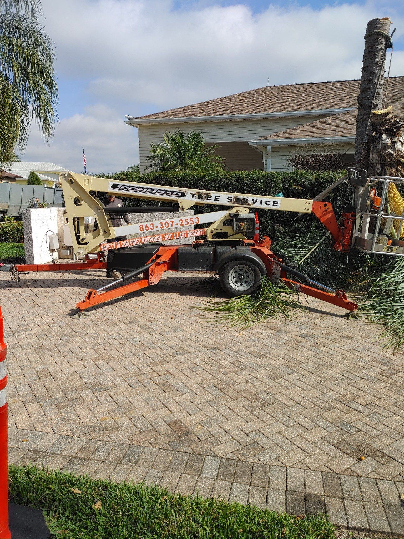 A cherry picker reaching a tree. Orange and beige construction equipment on a brick paved area next to a house.