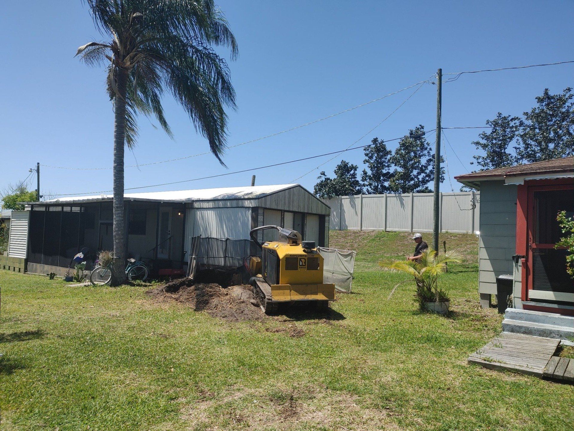 A small yellow construction vehicle works on a grassy lot near a mobile home and a small house on a sunny day.