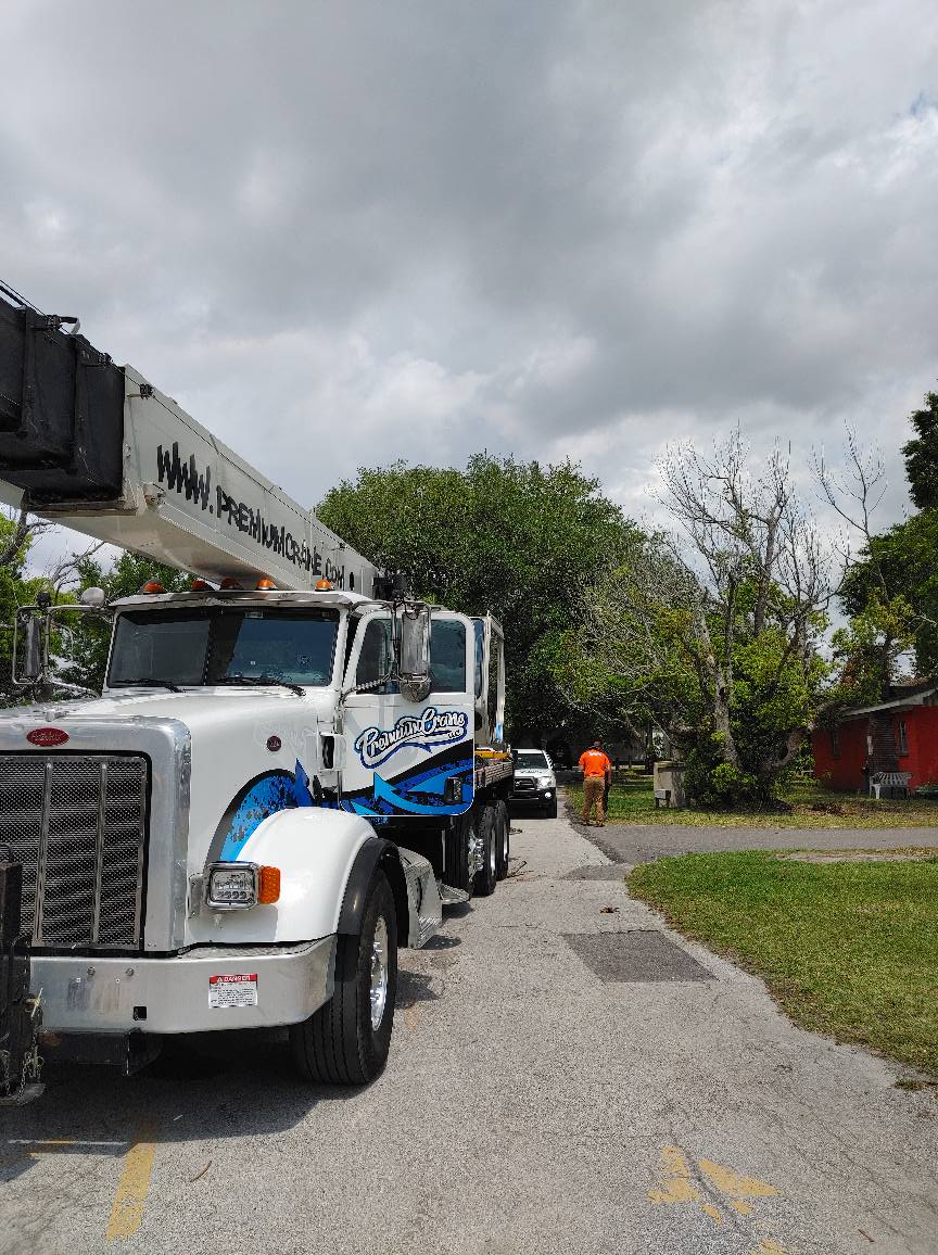 A white crane truck parked on asphalt next to a tree, an orange-clad worker, and a car. Cloudy sky.
