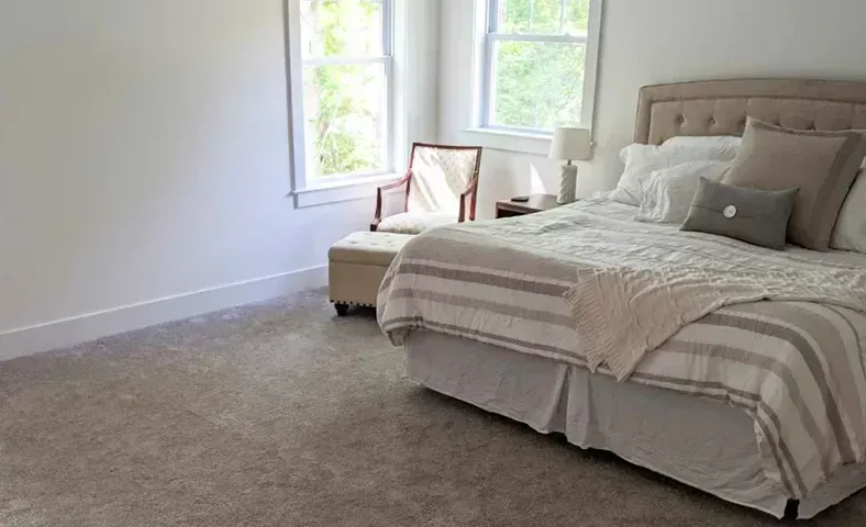 A neutral-toned bedroom with a bed, chair, and windows. Light-colored walls, bedding, and a carpet create a bright and airy space.